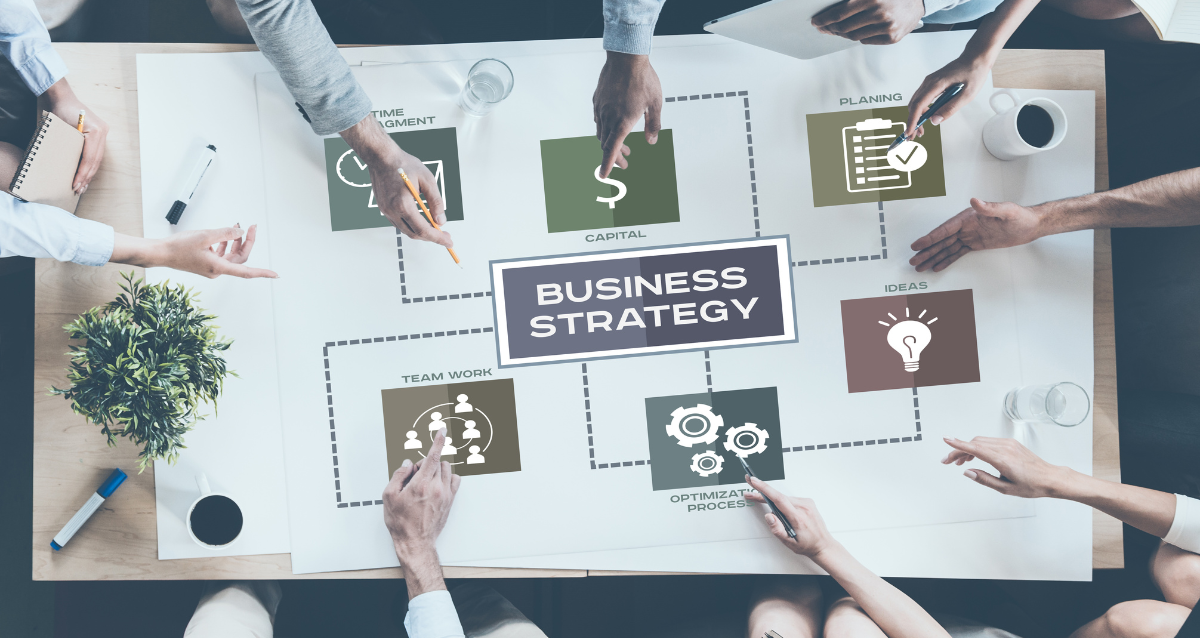 Wide angle photo of a table with multiple hands pointing at different aspects of a business (planning, team work, capital etc). 